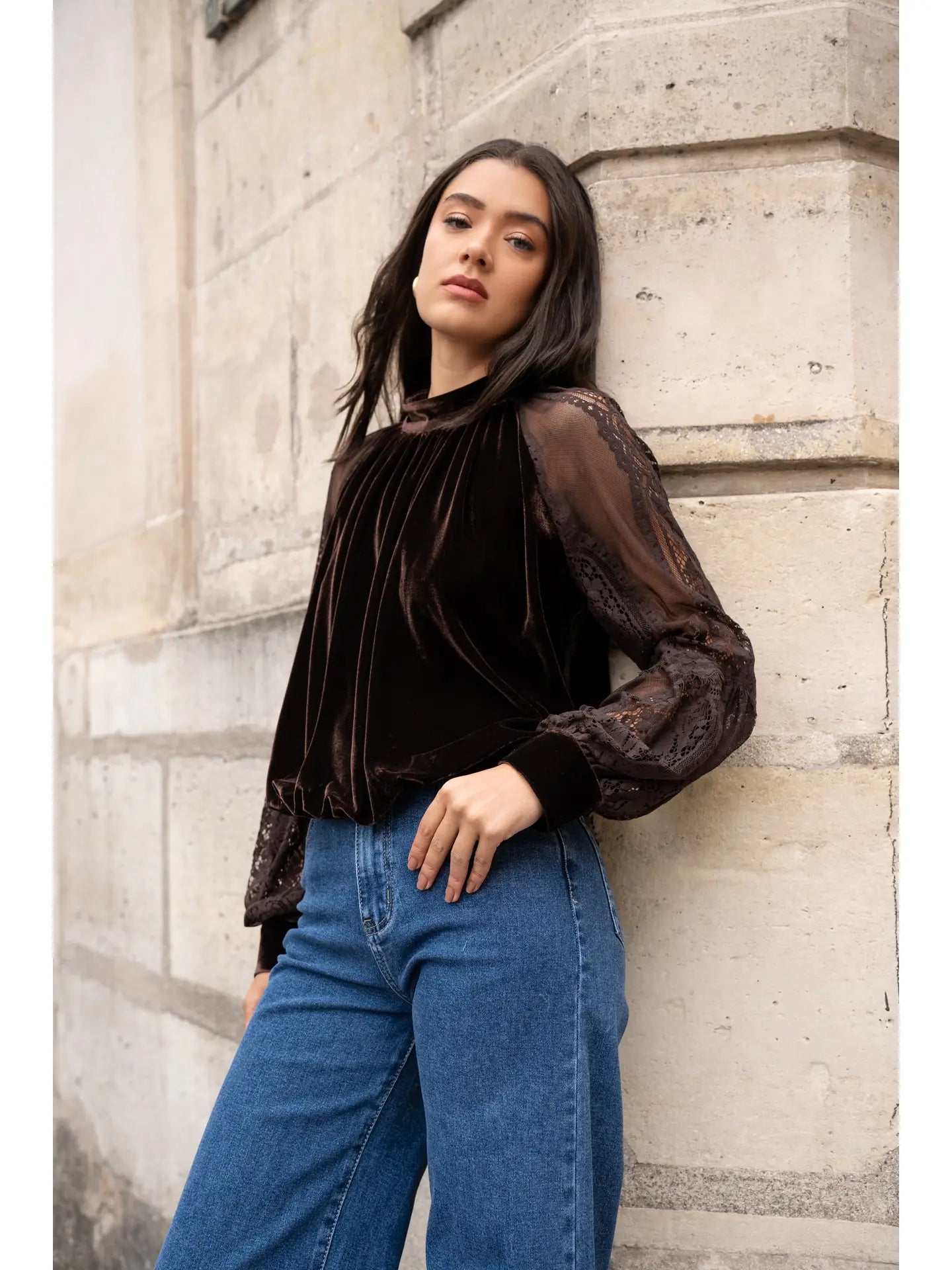 Woman wearing a dark blouse with sheer sleeves and blue jeans against a stone wall.