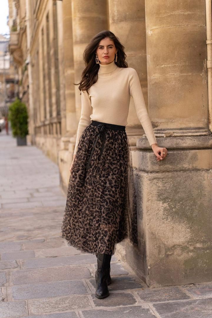 Woman in beige sweater and leopard print skirt standing against a stone wall.