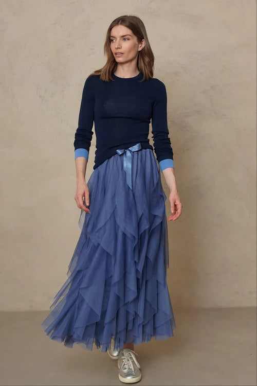 A woman modeling a dark navy long-sleeve knit top and a layered blue tulle midi skirt, paired with white sneakers, set against a neutral textured beige studio backdrop.