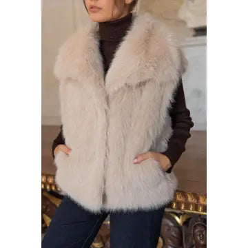 A woman wearing a fluffy light beige faux fur vest over a dark brown turtleneck sweater and dark blue jeans, standing in an indoor setting with a decorative gold chair visible in the background.