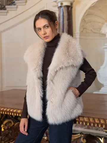 A woman wearing a light beige faux fur vest over a dark brown turtleneck sweater and dark blue jeans, posing in an elegant indoor setting with classical columns and a staircase.