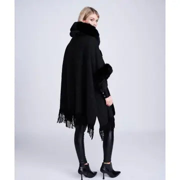 A model wearing a black fringed poncho with a plush collar, styled with black leggings and black high-heeled boots against a light gray studio background.