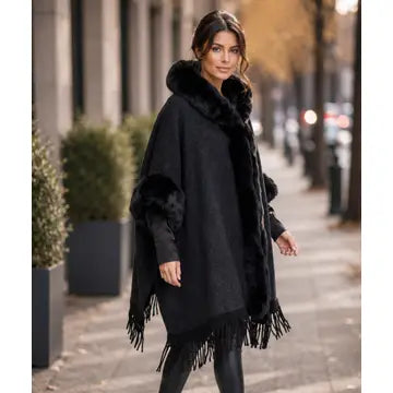 A woman wearing a black fringed poncho with a plush faux-fur collar, standing on a city sidewalk.