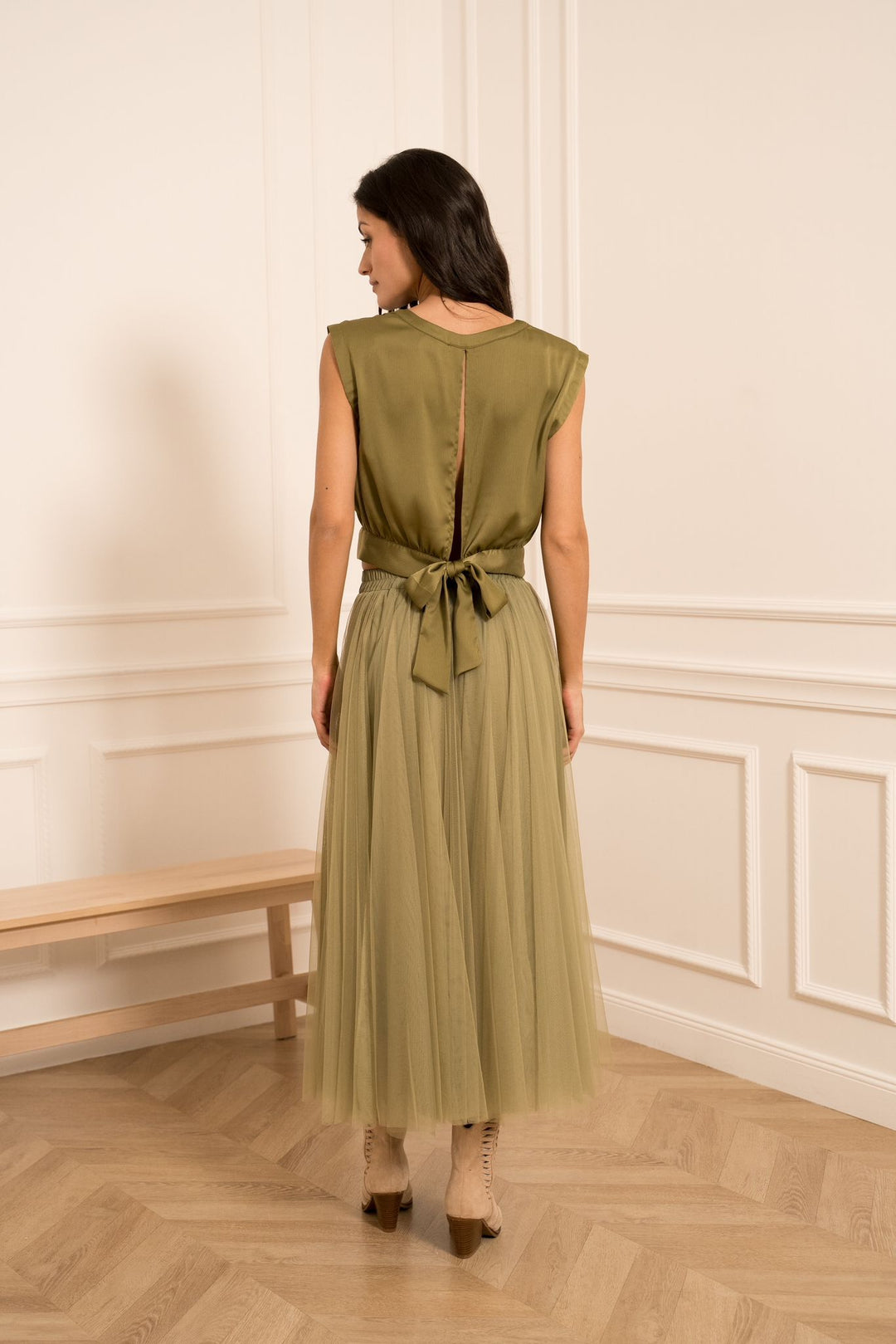 Woman wearing a green dress standing in a room with wooden flooring and white walls.