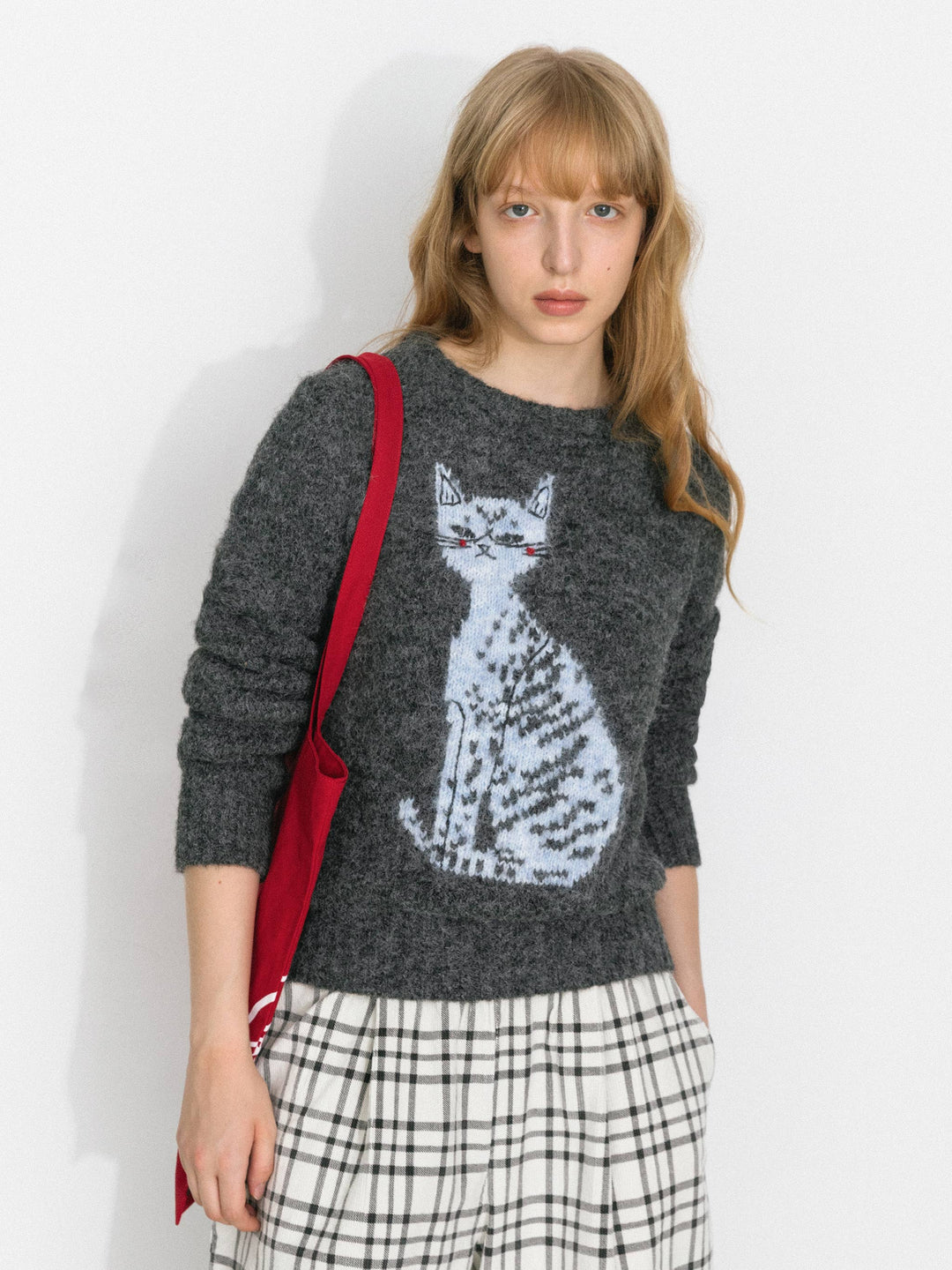 Woman wearing a sweater with a cat design, holding a red bag against a white background