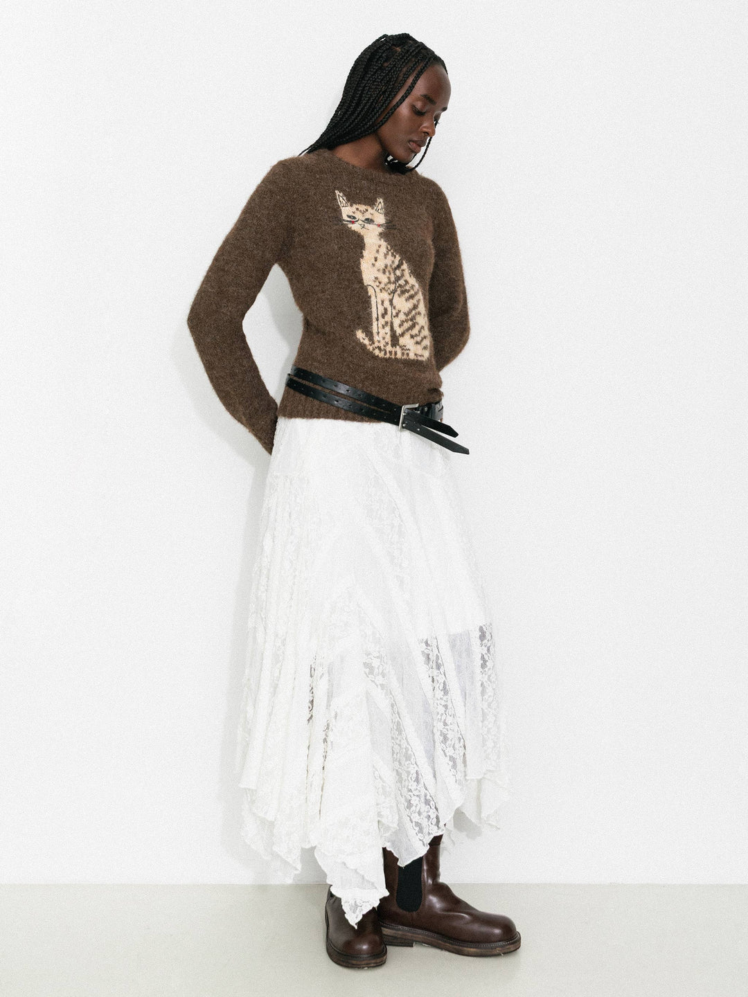 Woman wearing a brown sweater with a cat design, white skirt, and brown boots on a white background