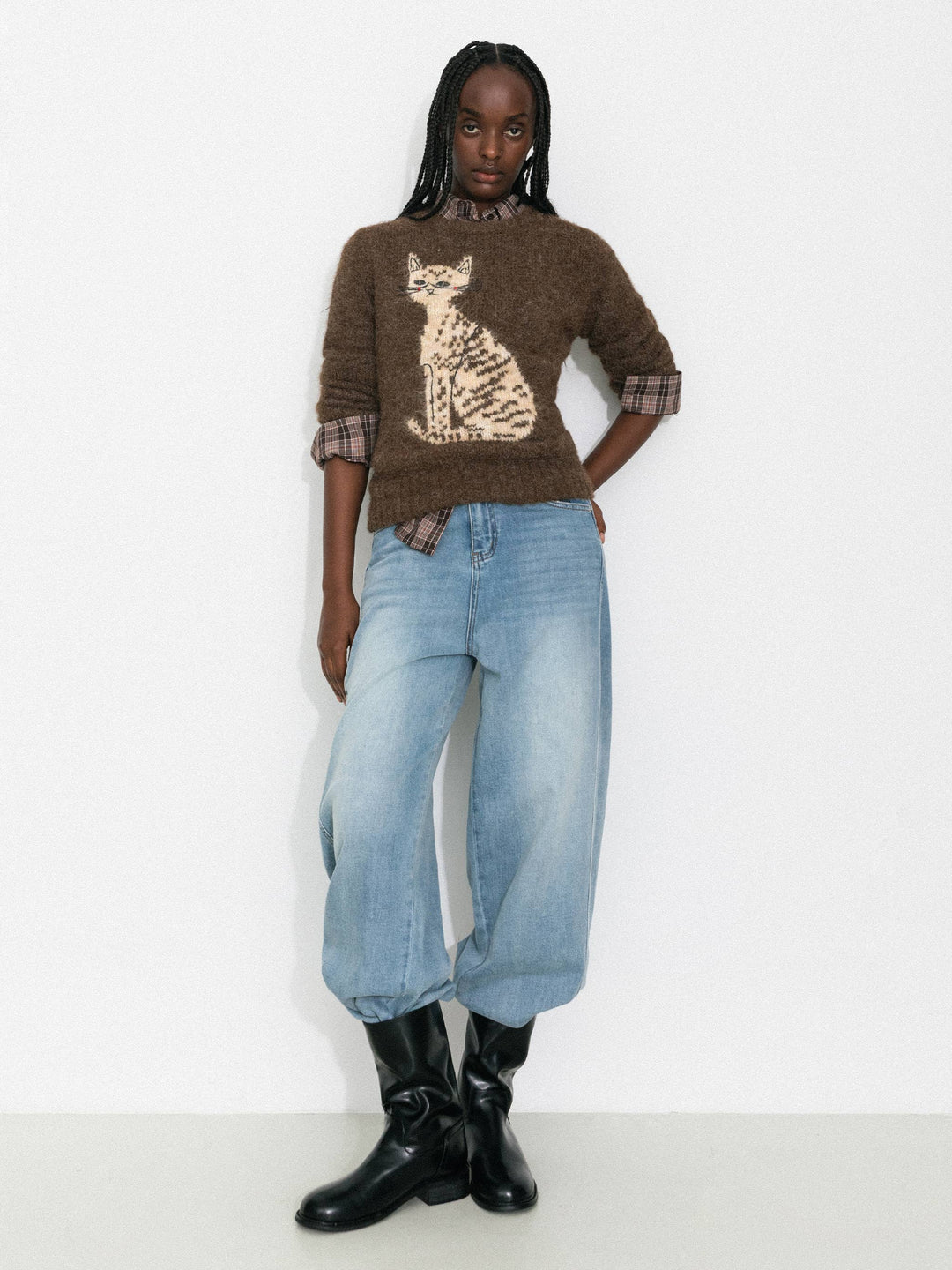 Person wearing a brown sweater with a cat design, blue jeans, and black boots on a white background