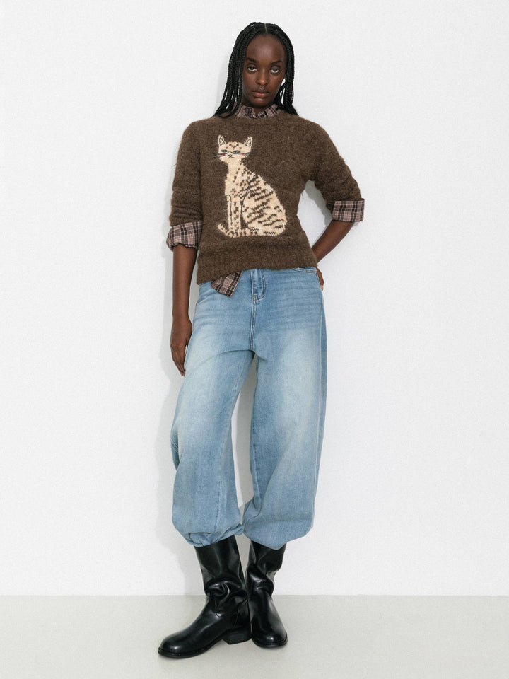 Person wearing a brown sweater with a cat design, blue jeans, and black boots on a white background