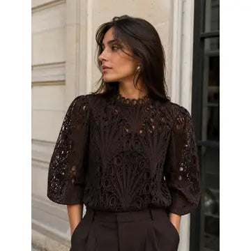 A woman wearing a dark brown lace blouse with puff sleeves and a high neckline, paired with dark trousers, standing outdoors against a light-colored building wall.