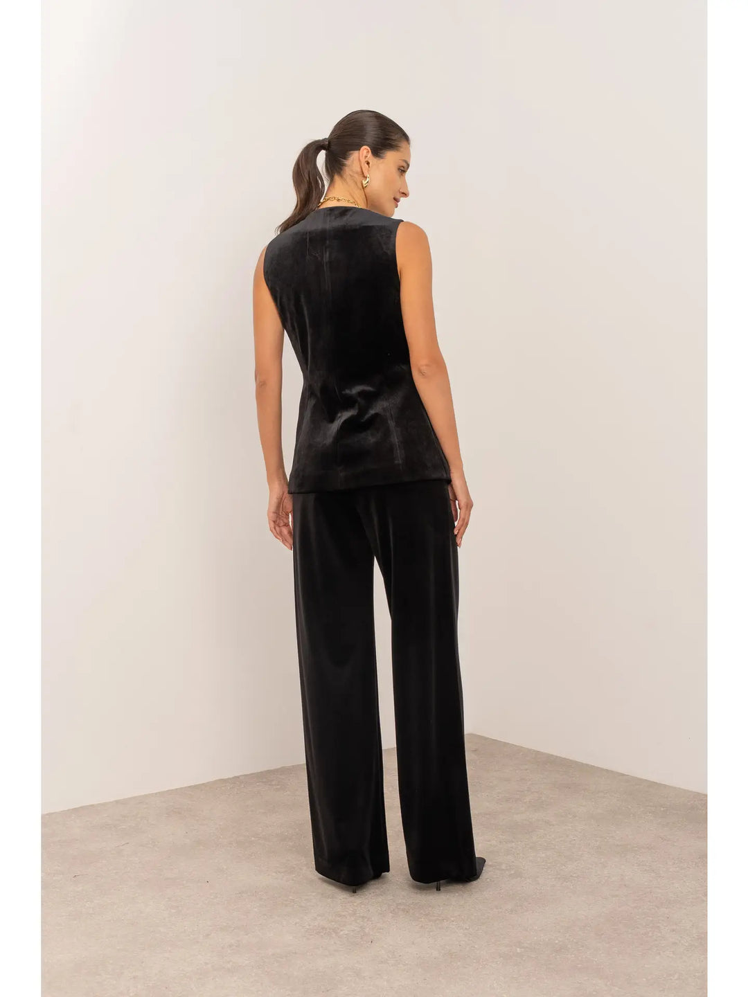 Woman wearing a black sleeveless top and wide-leg pants against a white wall.