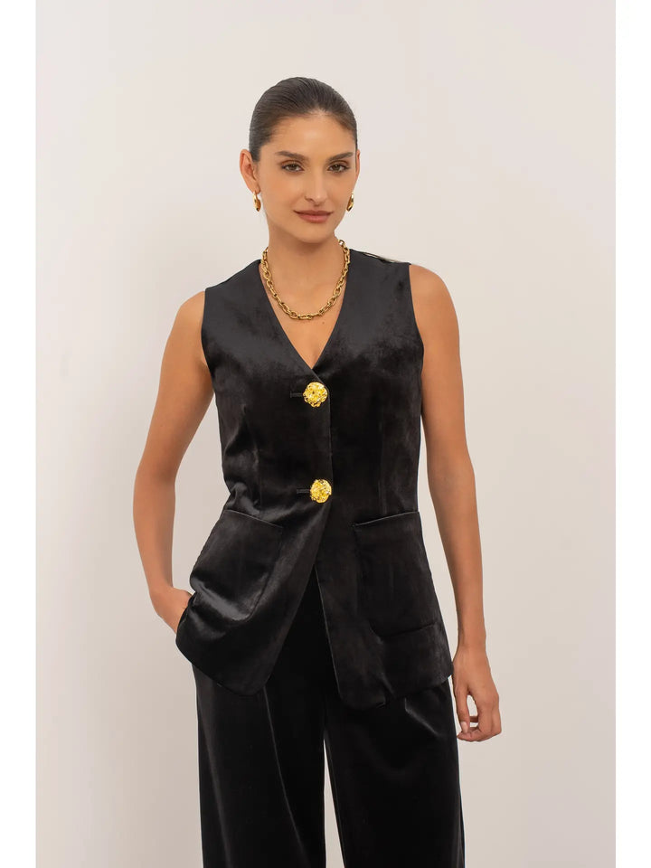 Woman wearing a black sleeveless top with gold buttons and black pants against a white background