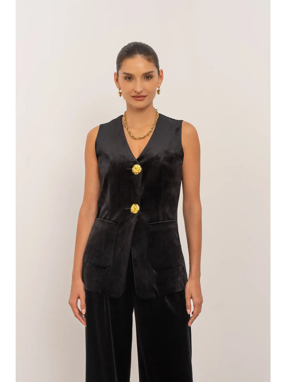 Woman wearing a black sleeveless blazer with gold buttons on a plain background