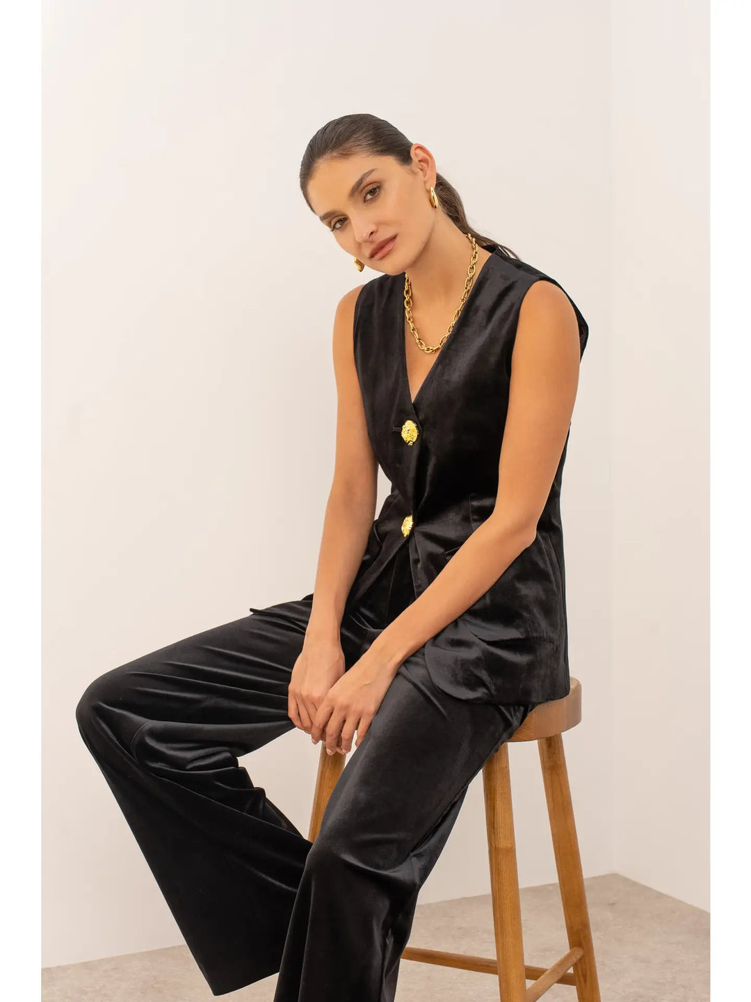 Woman wearing a black sleeveless top and pants sitting on a wooden stool against a white wall.