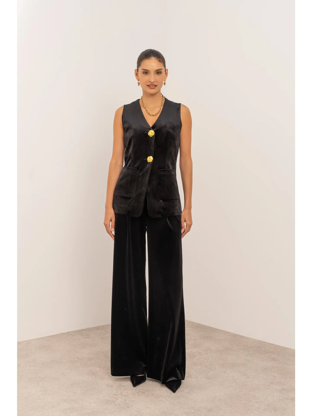 Woman wearing a black sleeveless blazer with gold buttons and wide-leg pants on a plain background