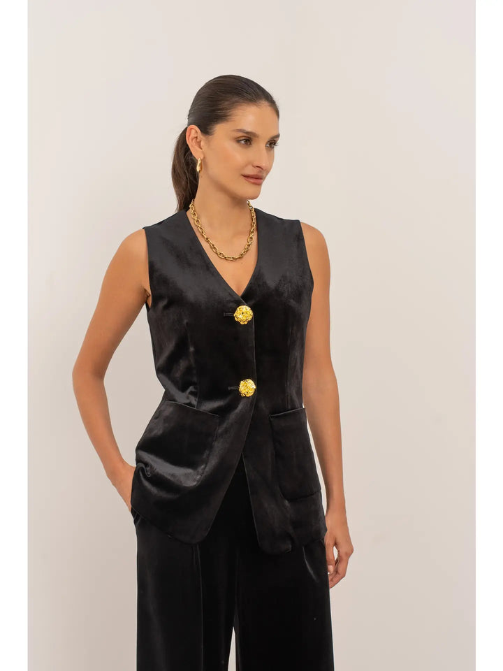 Woman wearing a black sleeveless top with gold buttons on a plain background