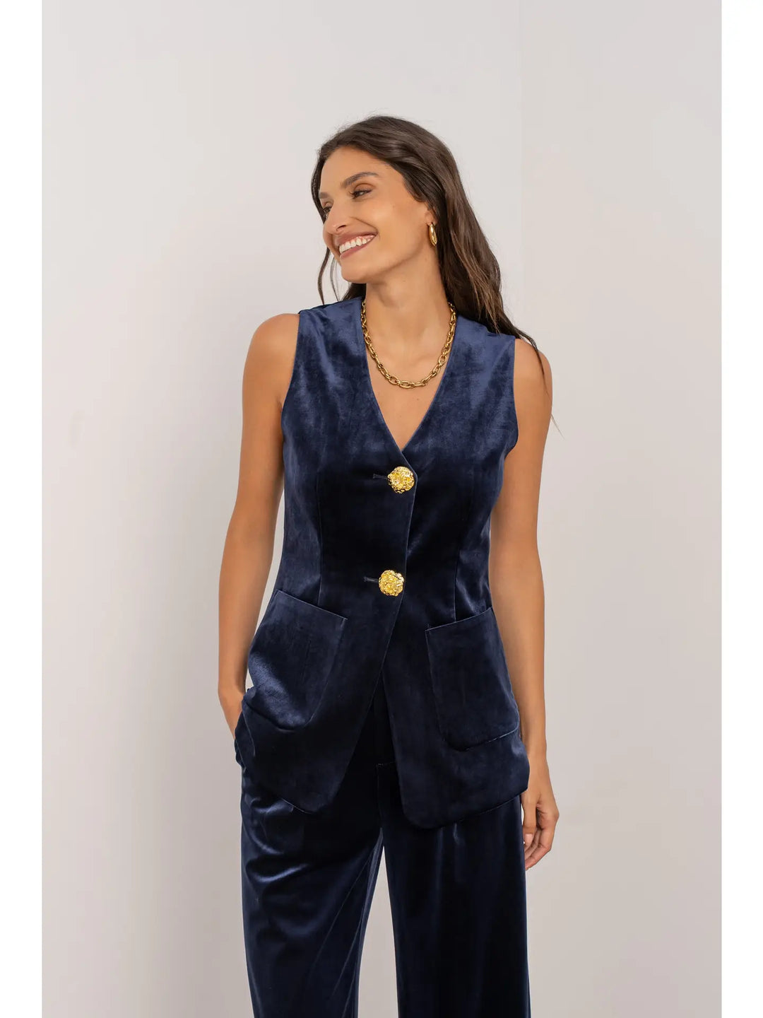 Woman wearing a navy blue vest and pants set with gold buttons on a light gray background