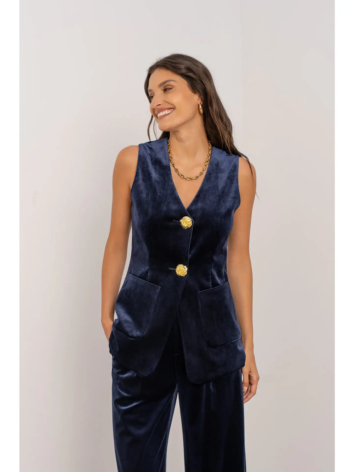 Woman wearing a navy blue vest and pants set with gold buttons on a light gray background