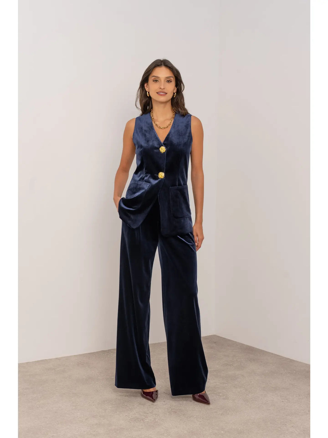 Woman wearing a navy velvet jumpsuit with gold buttons against a white background