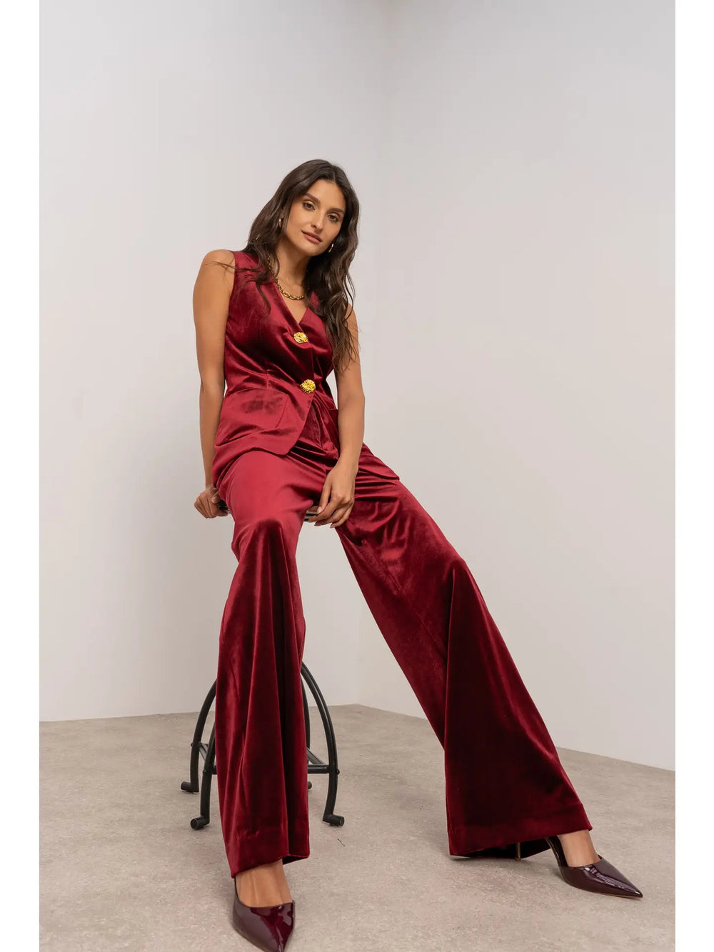 Woman wearing a burgundy velvet jumpsuit sitting on a stool against a plain background