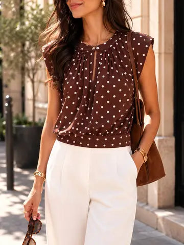 Woman wearing a brown polka dot top and white pants, holding a brown handbag.