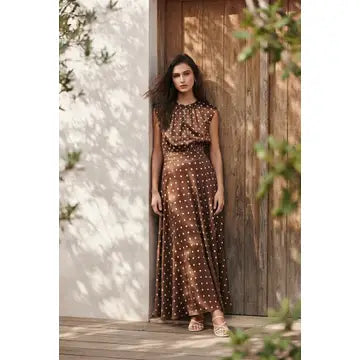 A woman stands outdoors in front of a rustic wooden door, wearing a sleeveless brown maxi dress with a white polka-dot pattern and a gathered, ruffled bodice.