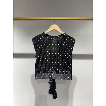A black sleeveless blouse with white polka dots, hanging on a wooden hanger against a light gray wall.