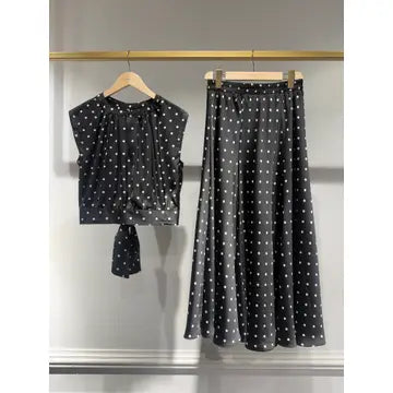 A two-piece women's outfit set featuring a short-sleeve tie-front top and a matching midi skirt, both in black with a white polka dot pattern, hanging on wooden hangers against a light gray wall.