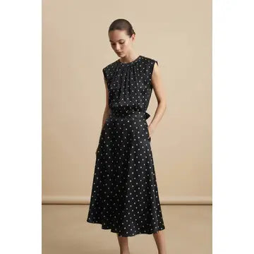 A woman modeling a sleeveless black midi dress with a white polka dot pattern, featuring a gathered high neckline and a flared skirt, set against a plain beige studio background.