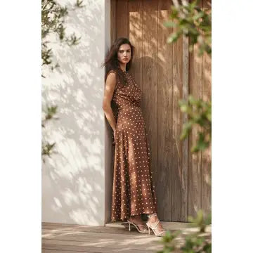A woman stands outdoors against a wooden door and white wall, wearing a sleeveless brown maxi dress with small white polka dots and nude high-heeled sandals.