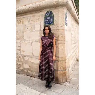 A woman stands outdoors against a stone wall corner wearing a sleeveless dark purple midi dress with white polka dots and black ankle boots. A blue street sign is visible on the wall.