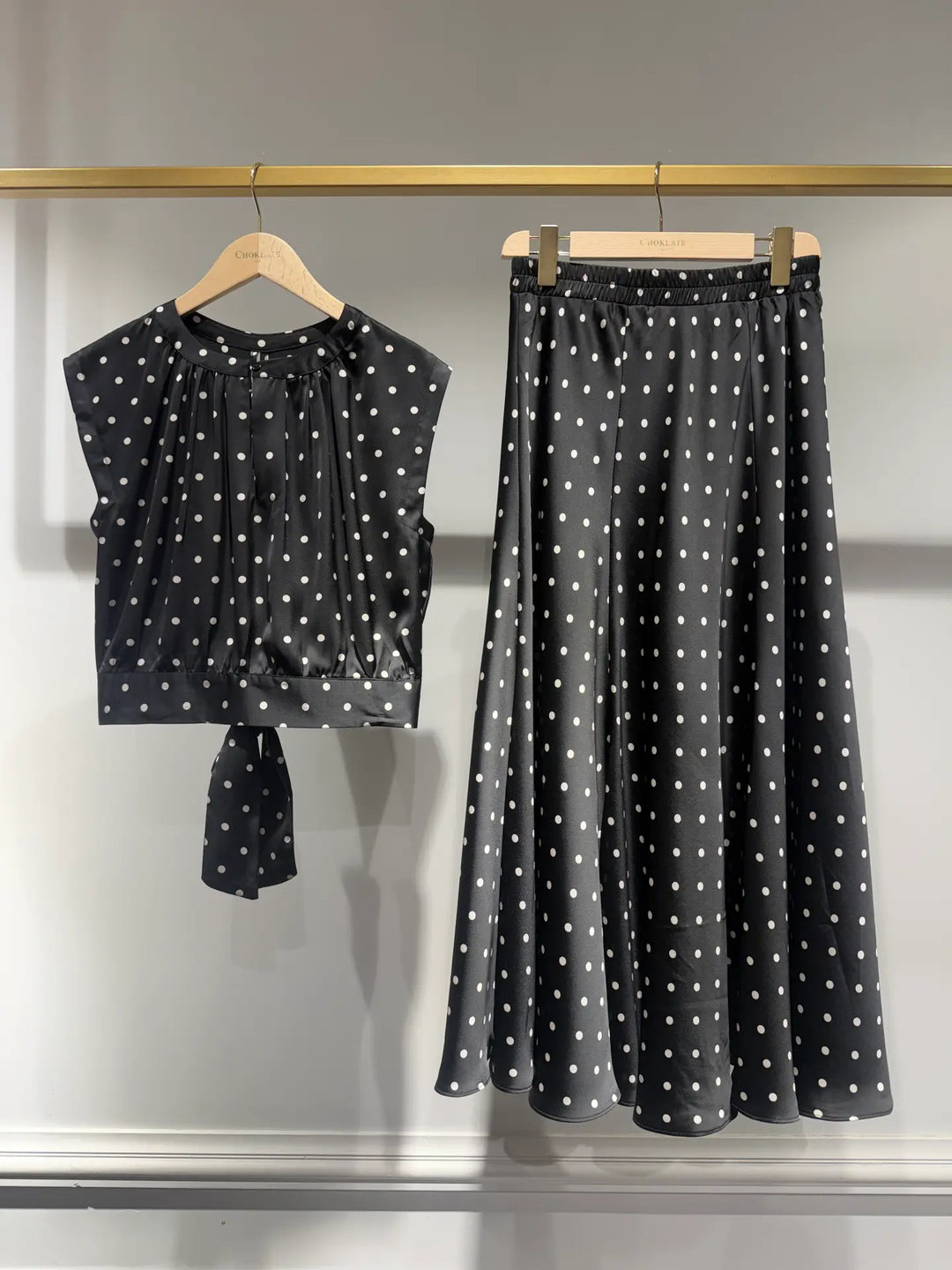A two-piece women's outfit set featuring a sleeveless black top with white polka dots and a matching high-waisted A-line skirt, both hanging on wooden hangers against a light gray wall.