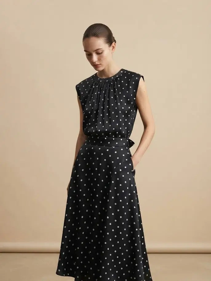A woman modeling a sleeveless black midi dress with a white polka dot pattern, standing against a solid beige studio background.