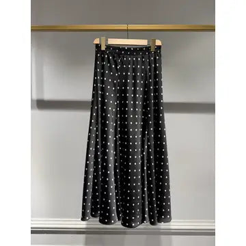 A black midi skirt with a white polka dot pattern, hanging on a wooden hanger from a gold clothing rack against a gray wall.