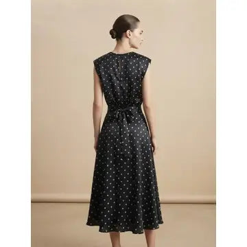 A woman wearing a sleeveless black midi dress with a white polka dot pattern, shown from the back against a solid beige studio background.