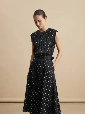 A woman modeling a sleeveless black midi dress with a white polka dot pattern, standing against a solid beige studio background.