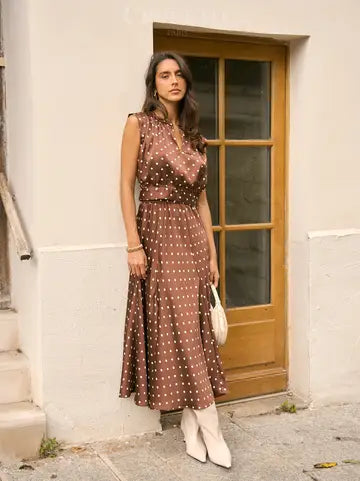 A woman stands outdoors in front of a cream-colored wall and wooden door, wearing a sleeveless brown midi dress with a white polka-dot pattern and white pointed-toe boots.