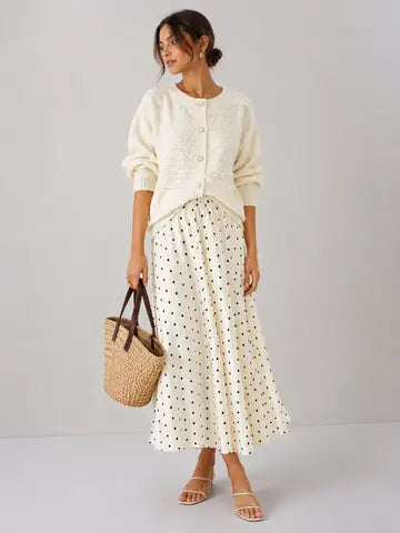Woman wearing a white cardigan and polka dot skirt holding a straw bag on a plain background