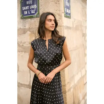 A woman wearing a black sleeveless dress with white polka dots stands against a stone wall with blue street signs.