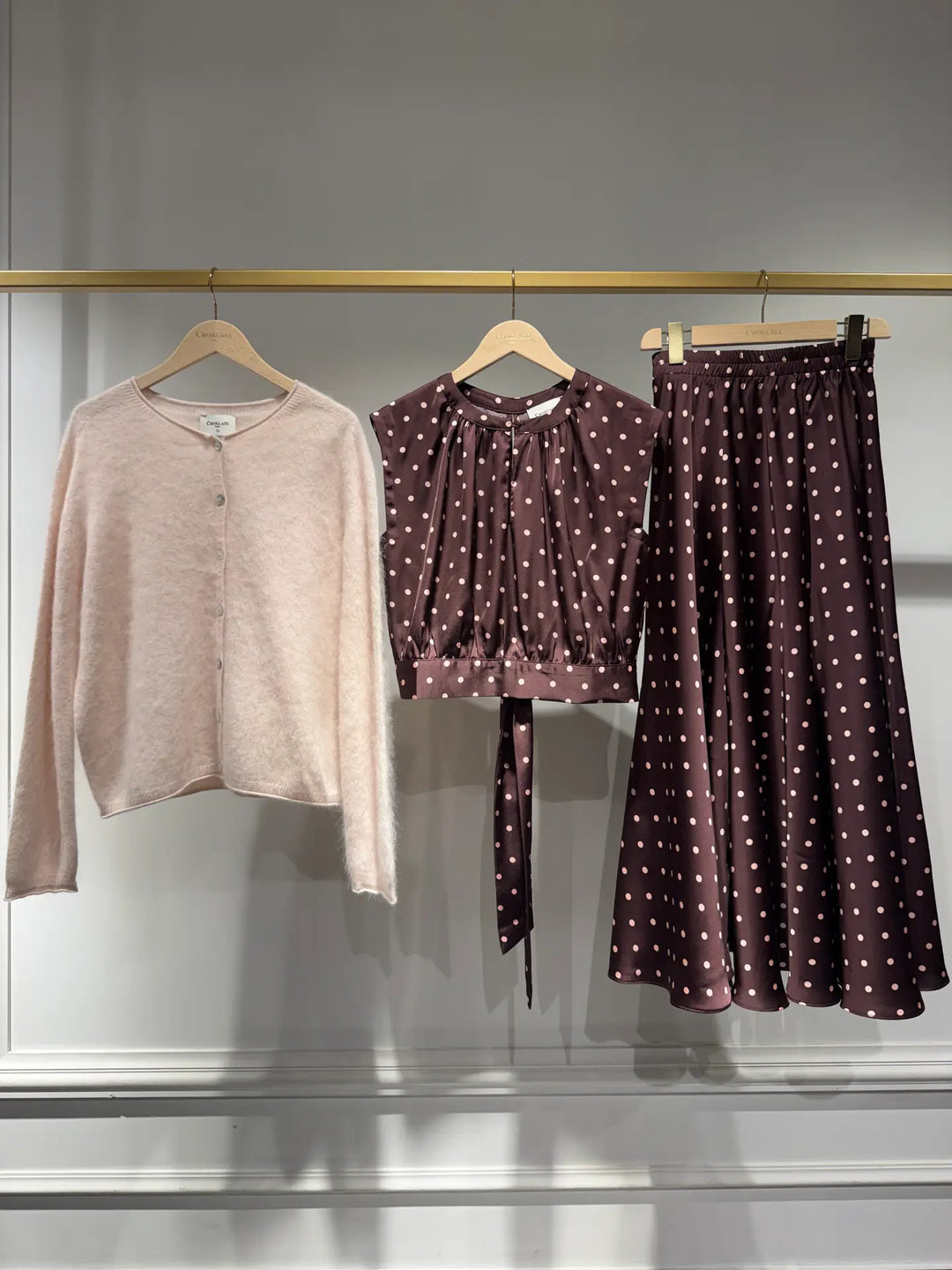 Three women's clothing items hanging on wooden hangers from a gold rail against a light gray wall: a light blush pink cardigan, a dark plum blouse with white polka dots and a tie detail, and a matching dark plum polka dot midi skirt.