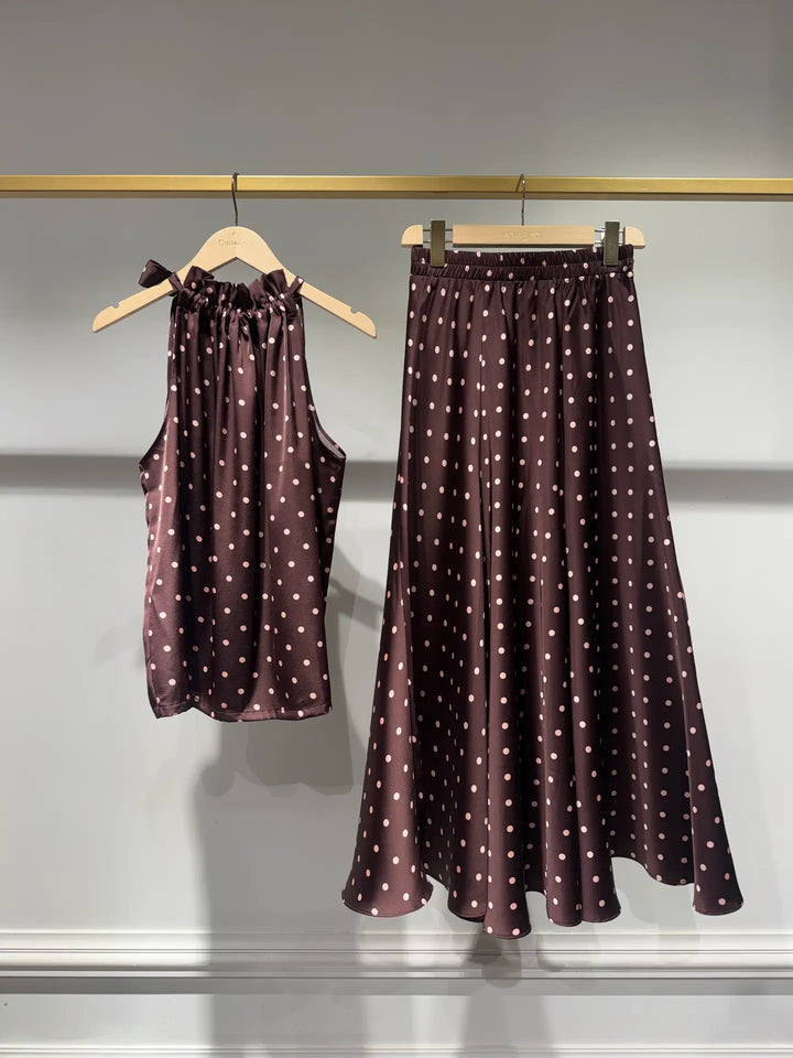 A matching two-piece clothing set consisting of a sleeveless halter top and a high-waisted A-line skirt, both in a dark brown satin fabric with a white polka dot pattern, hanging on wooden hangers against a light gray wall.