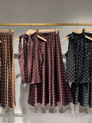 Three matching sleeveless top and skirt sets hanging on a gold clothing rack against a light gray wall. The center set is a deep plum/burgundy color with white polka dots, flanked by brown and black versions of the same pattern.
