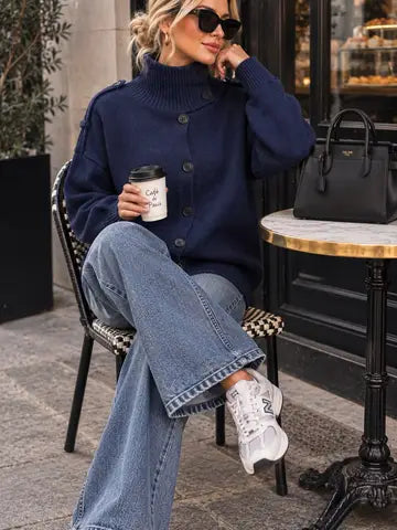 A woman sits on a chair at an outdoor cafe table wearing a navy blue button-up knit sweater, light-wash wide-leg jeans, and white sneakers. She holds a coffee cup next to a black handbag on a marble-topped table.