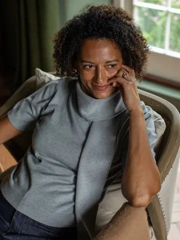 A woman with curly dark hair sits in a light-colored armchair indoors, wearing a gray short-sleeve mock-neck top and dark jeans. She is resting her head on her hand, with a window and green curtains visible in the background.