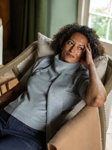 A woman with curly dark hair sits relaxed in a beige armchair indoors, wearing a light gray knit turtleneck sweater and dark blue jeans, with her hand resting on her head.