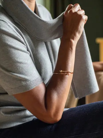 A close-up lifestyle shot of a person wearing a delicate gold bangle bracelet with a safety-pin style clasp, paired with a light gray knit sweater.