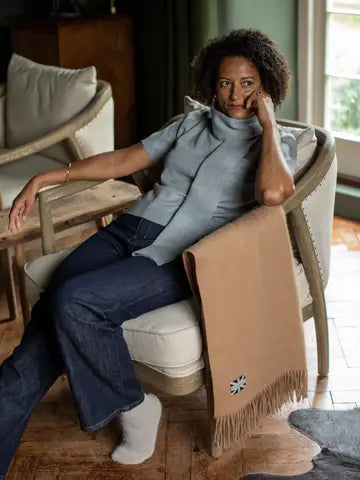 A woman sits in a light-colored armchair in a home interior, wearing a light blue turtleneck top and dark blue jeans. A tan fringed scarf with a small Union Jack logo is draped over the arm of the chair.