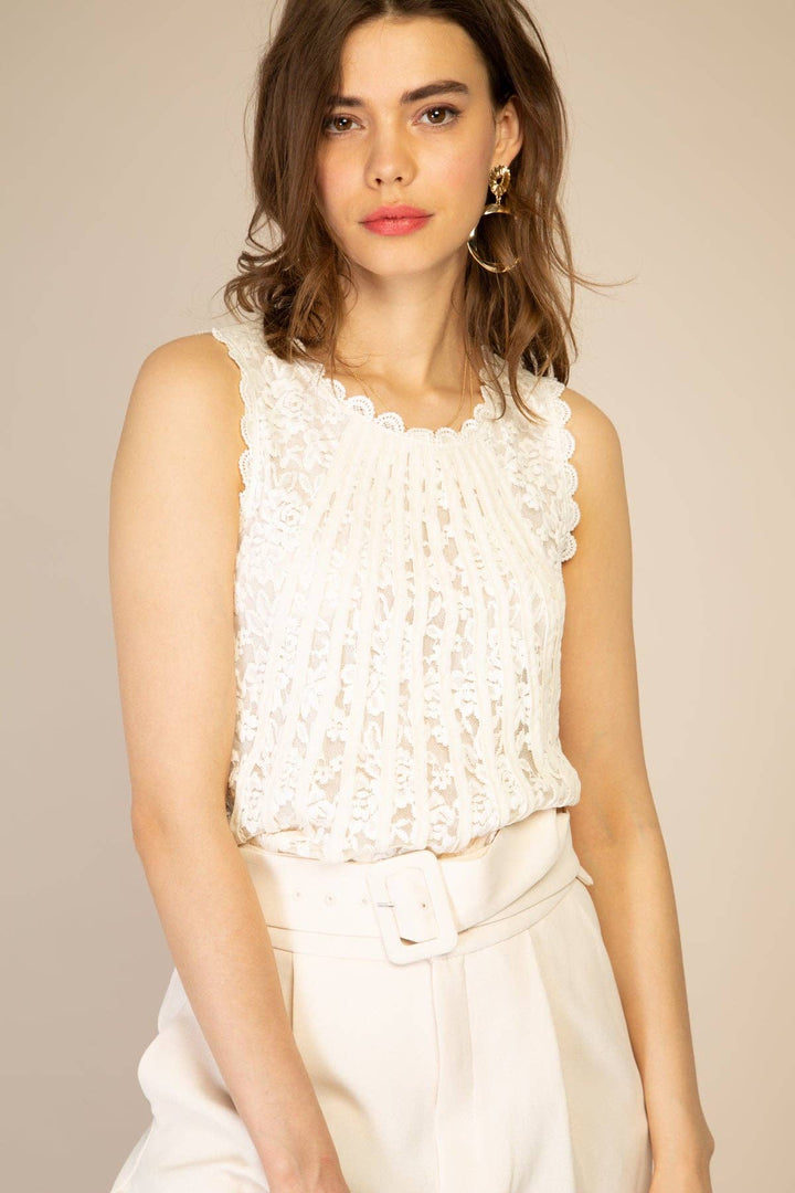 Woman wearing a white sleeveless top with lace details against a beige background