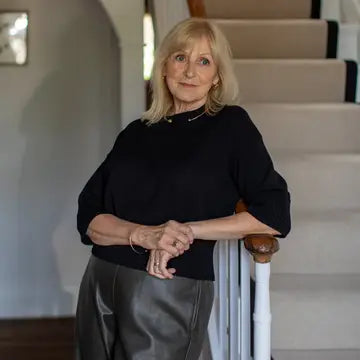 An older woman with blonde hair stands indoors near a staircase, wearing a black long-sleeve top and a dark gray skirt.