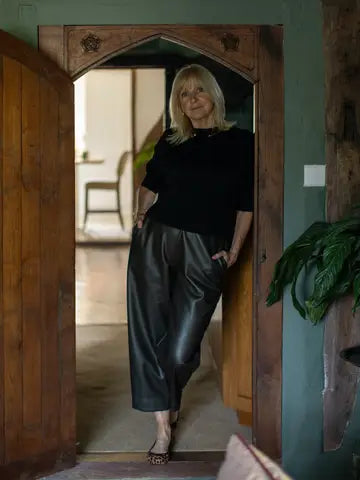 A woman with blonde hair stands in a doorway wearing a black long-sleeve top and shiny black wide-leg pants, with a home interior visible behind her.