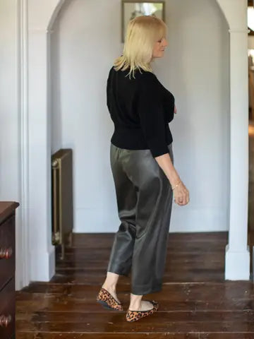 A blonde woman is shown from the back wearing a black three-quarter sleeve top, dark gray satin-like cropped pants, and leopard print flats, standing in a hallway with white walls and a dark wood floor.